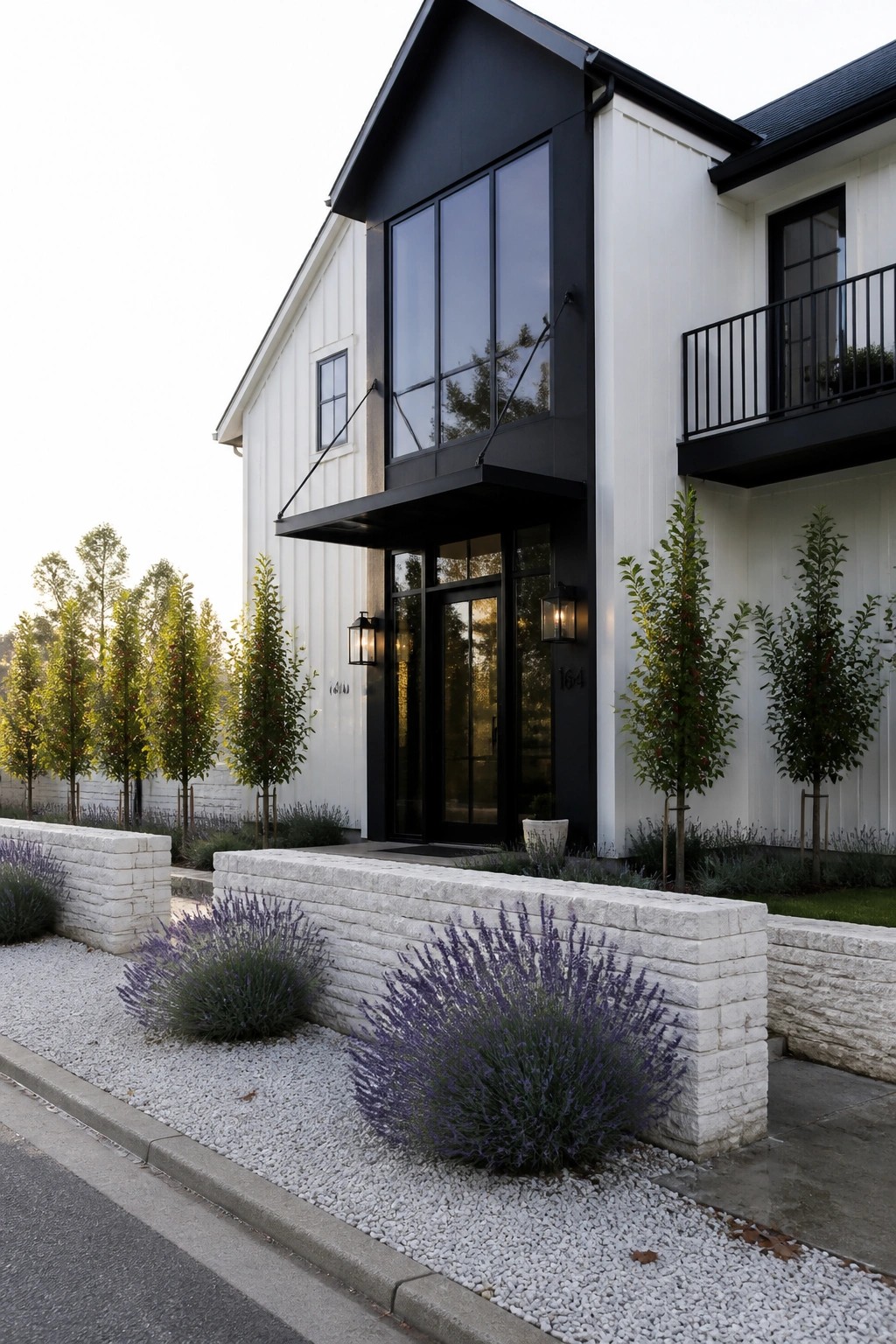A two-story white farmhouse with black trim around tall windows and the front entry, gravel landscaping, and lavender bushes in front.