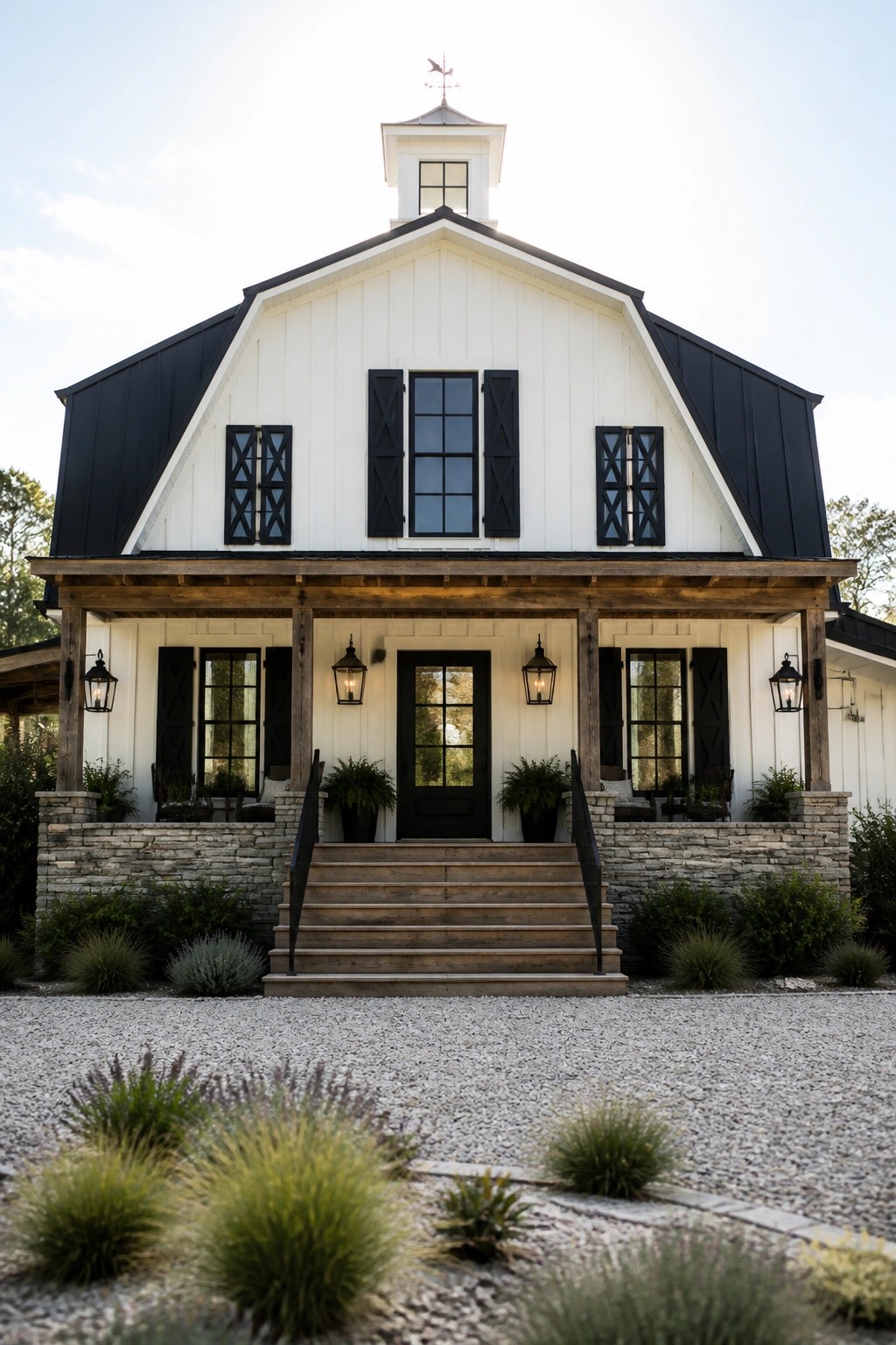 A white barn-style farmhouse with a black metal roof, black shutters, a black front door, wooden porch posts, and a stone foundation under a gravel driveway.