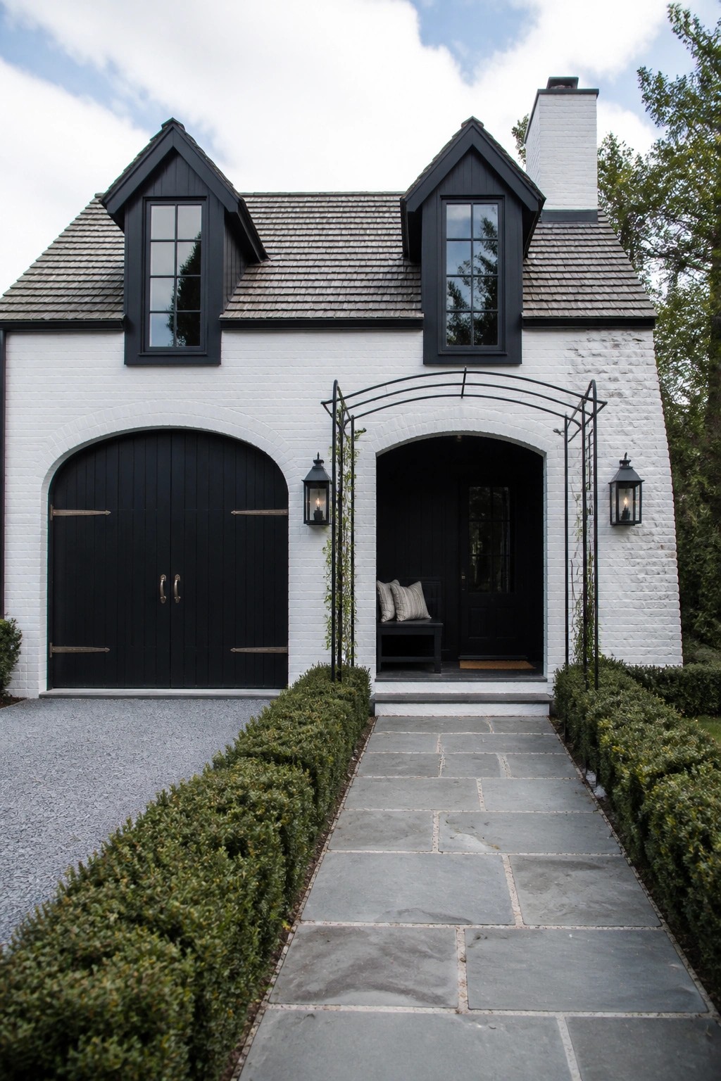 A white brick farmhouse exterior with black arched garage doors on the left, a black front door under an arched opening on the right, two black dormer windows, and a stone pathway bordered by hedges.