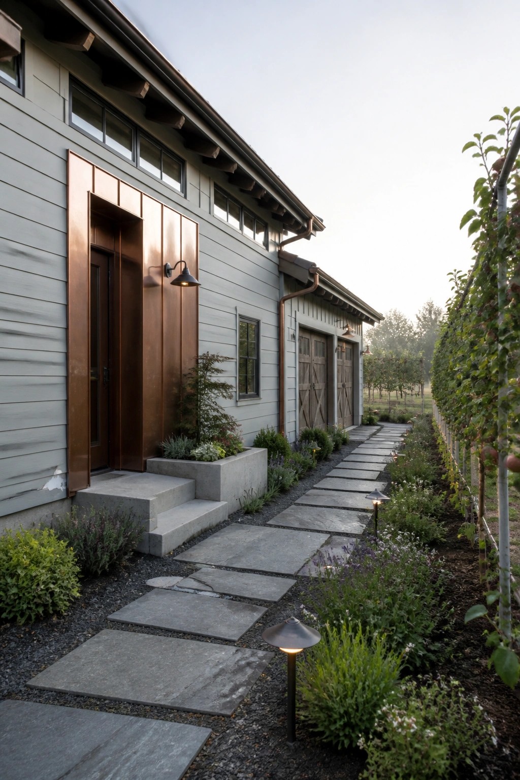 A side view of a farmhouse exterior showing gray horizontal siding, copper metal panels framing an entry door, two wooden barn-style doors further down the wall, and a stone pathway bordered by garden beds.