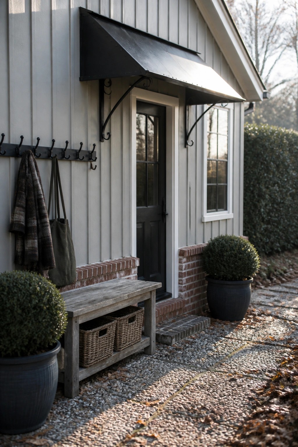 Gray vertical siding farmhouse exterior with black metal awning over black door, brick base, and potted shrubs beside a wooden bench.