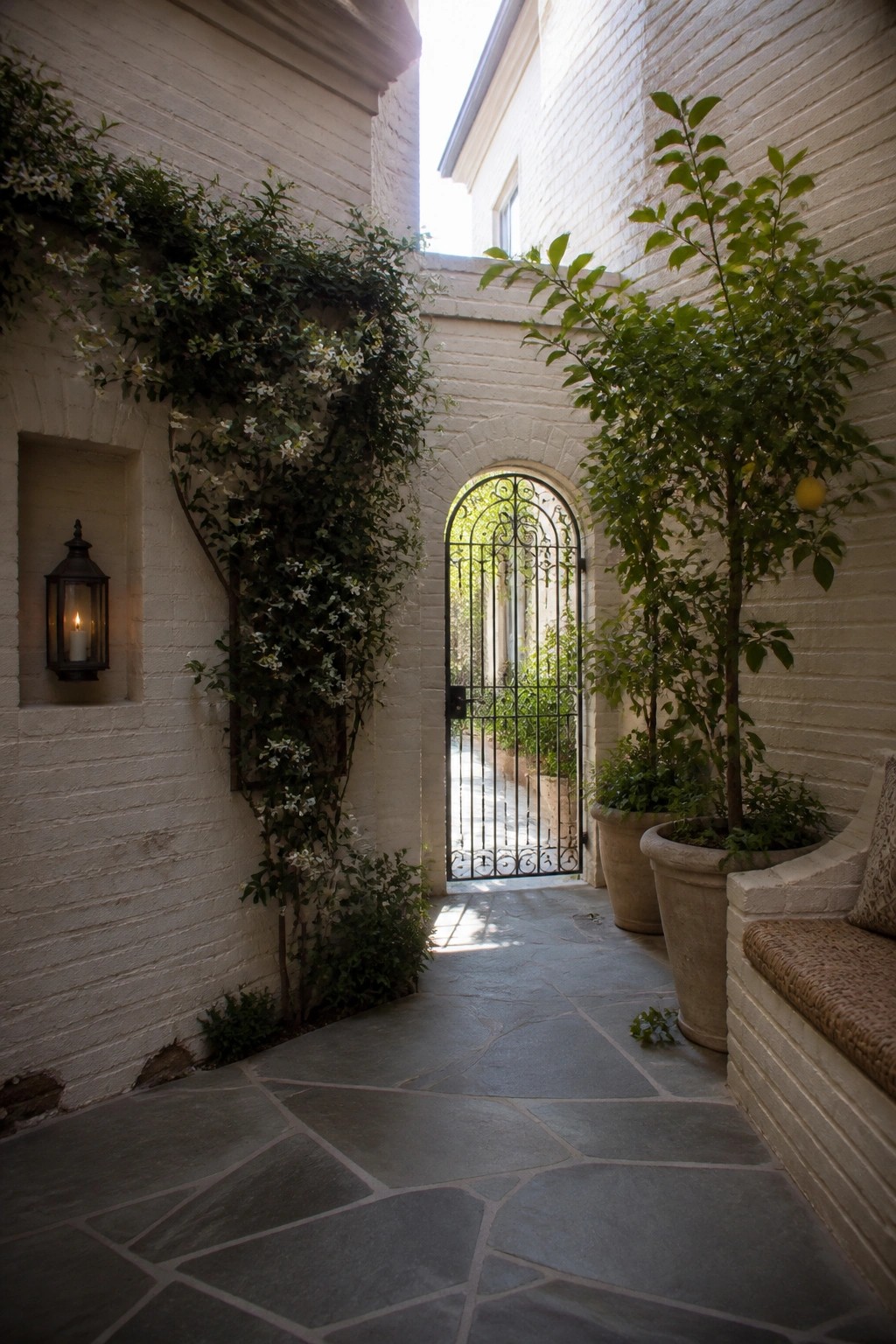 A narrow stone path runs between white brick walls with climbing vines, two large potted trees, a lit lantern niche, a built-in bench, and an arched metal gate at the far end.