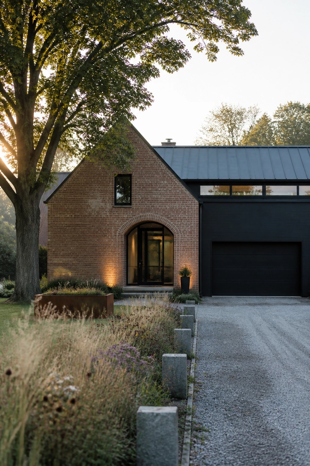 A red brick farmhouse exterior with a prominent arched entryway containing a black glass door, next to a large black garage door, set beside a gravel driveway and large tree.