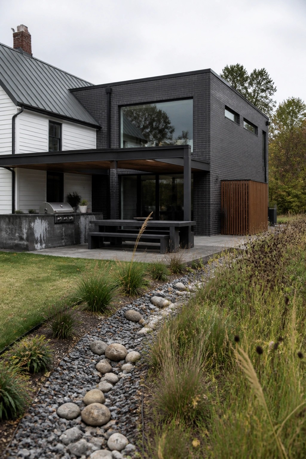 A modern black brick addition attached to a white farmhouse with a covered patio, outdoor table, and gravel landscaping in the foreground.