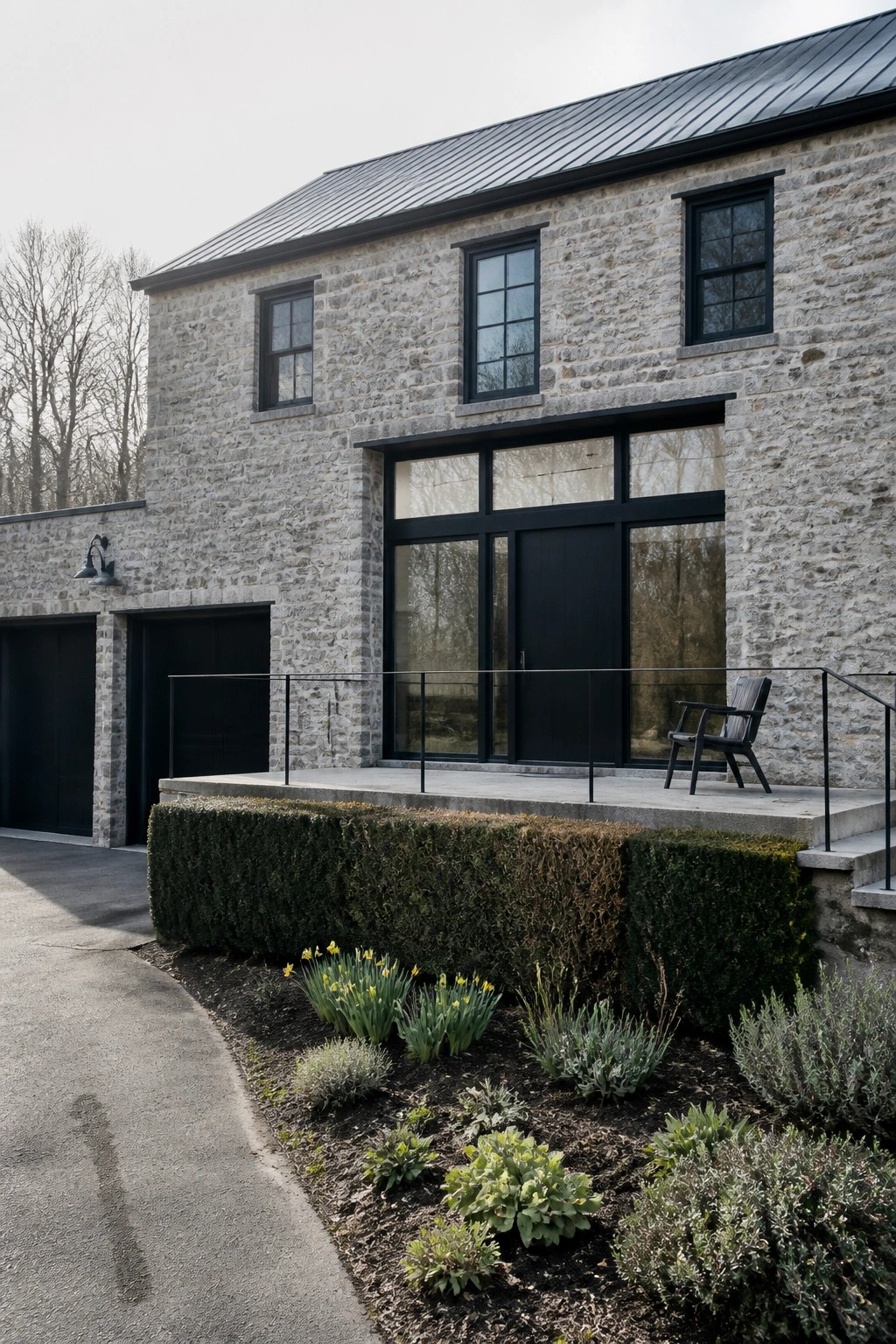 A stone farmhouse exterior featuring black-framed windows and doors, a raised concrete deck with a single chair, and low garden beds along the front.