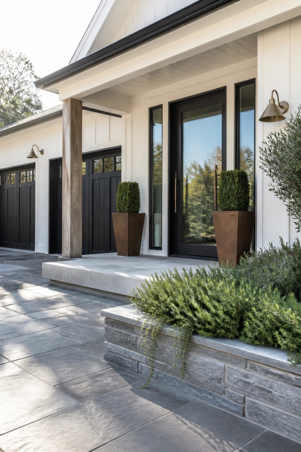 A front porch view of a white house with black doors and windows, wooden posts, tall planters, and a stone retaining wall with greenery.