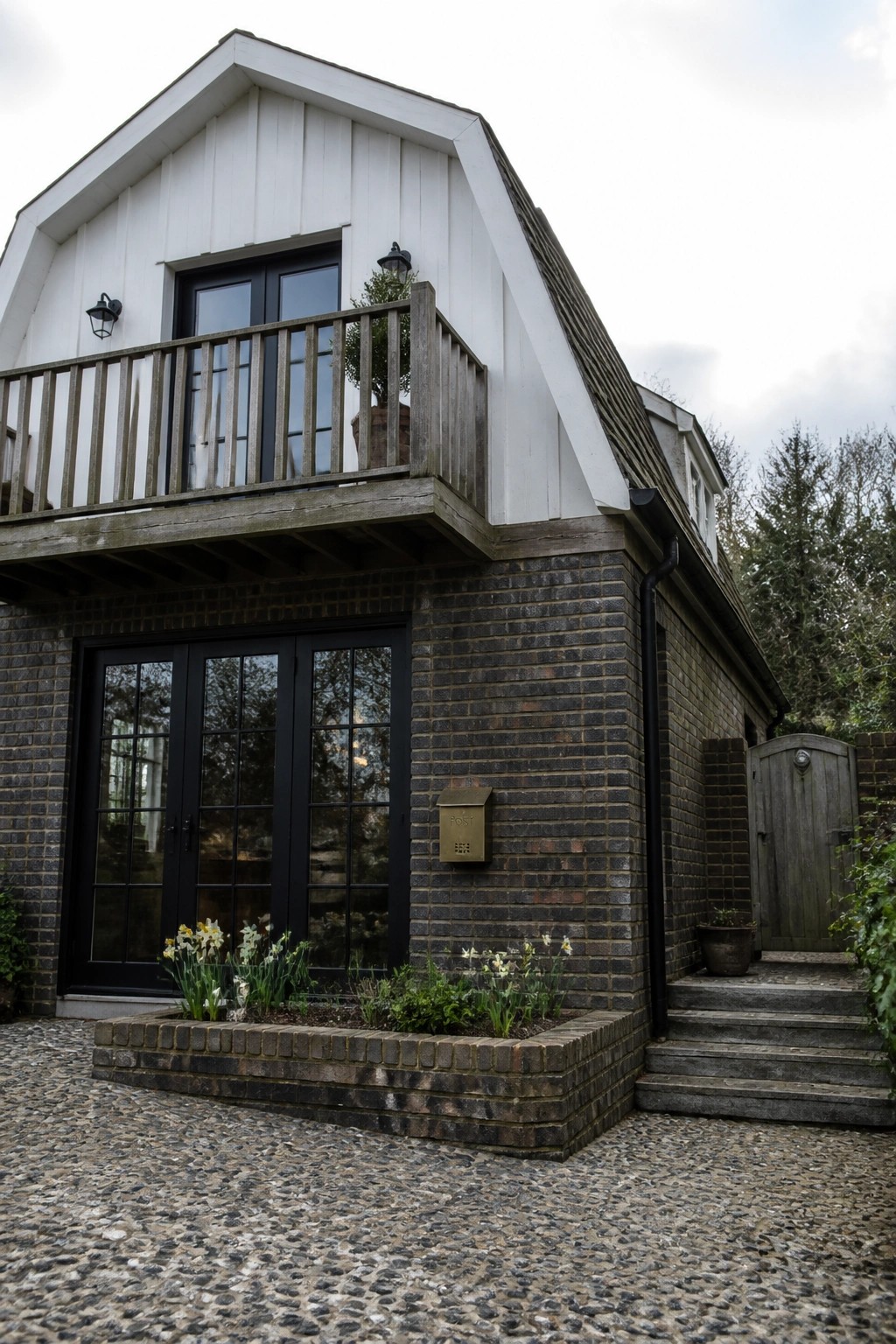 A two-story house with dark brick on the lower level, white vertical siding above, black-framed doors and windows, a wooden balcony, and a brick planter with flowers in front.