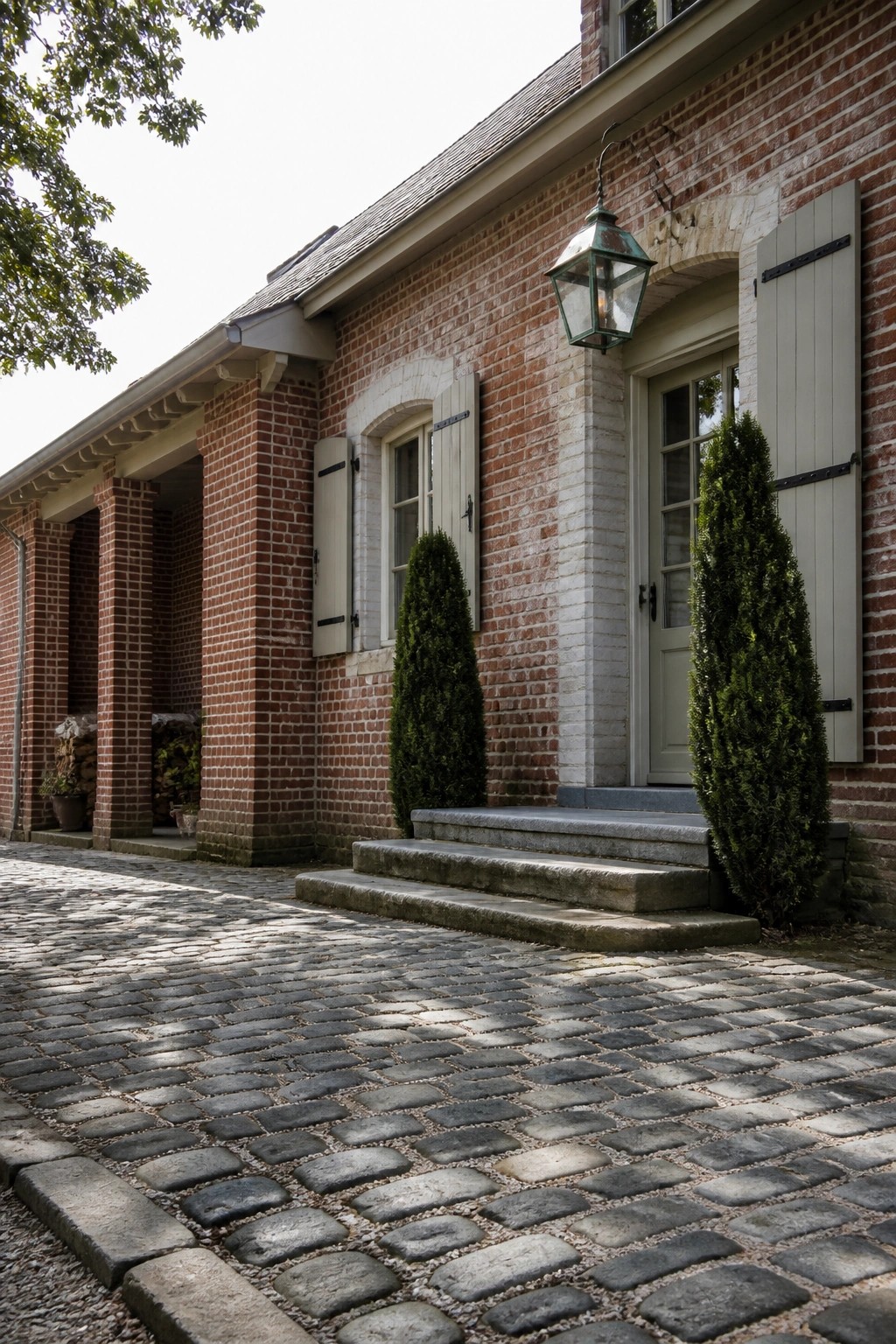 A red brick house exterior with light shutters, a green door, stone steps, a hanging lantern, and a cobblestone path in the foreground.