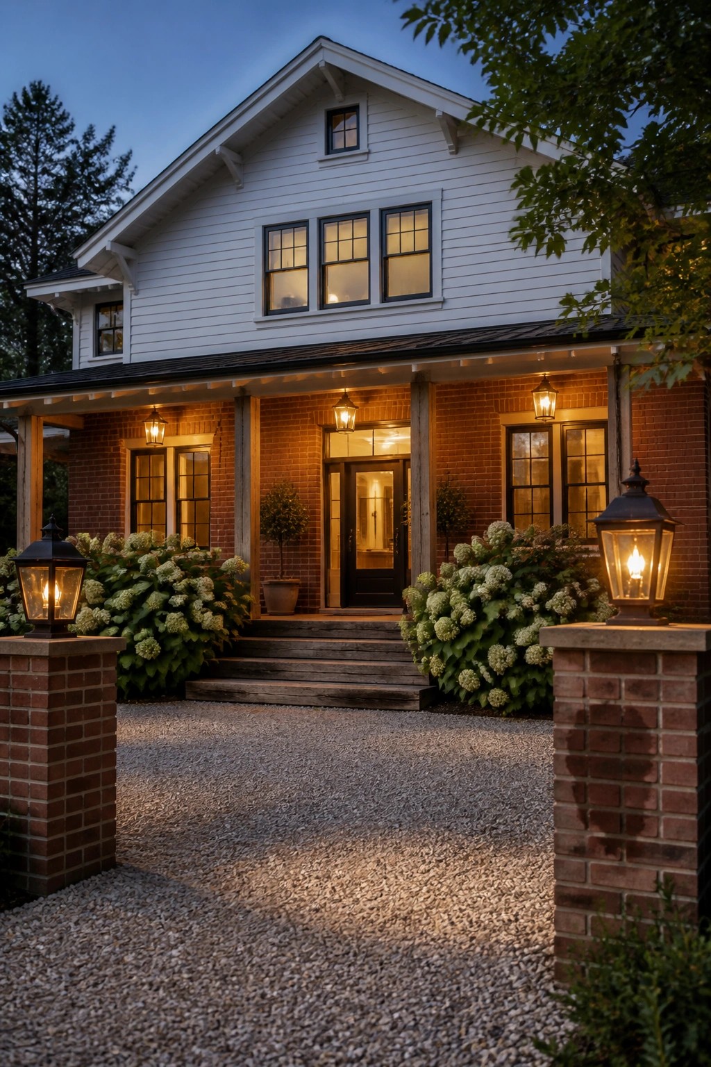 A two-story white farmhouse with red brick on the lower level, a covered front porch, multiple glowing lanterns, and a gravel driveway at dusk.