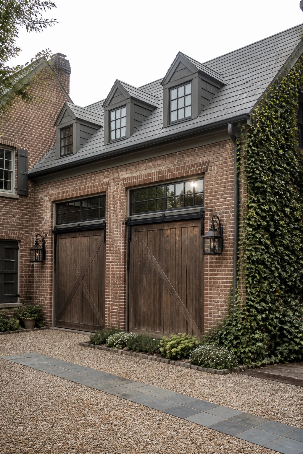 A brick house exterior featuring two large wooden garage doors with black hardware, dormer windows above, and ivy growing along the right side.