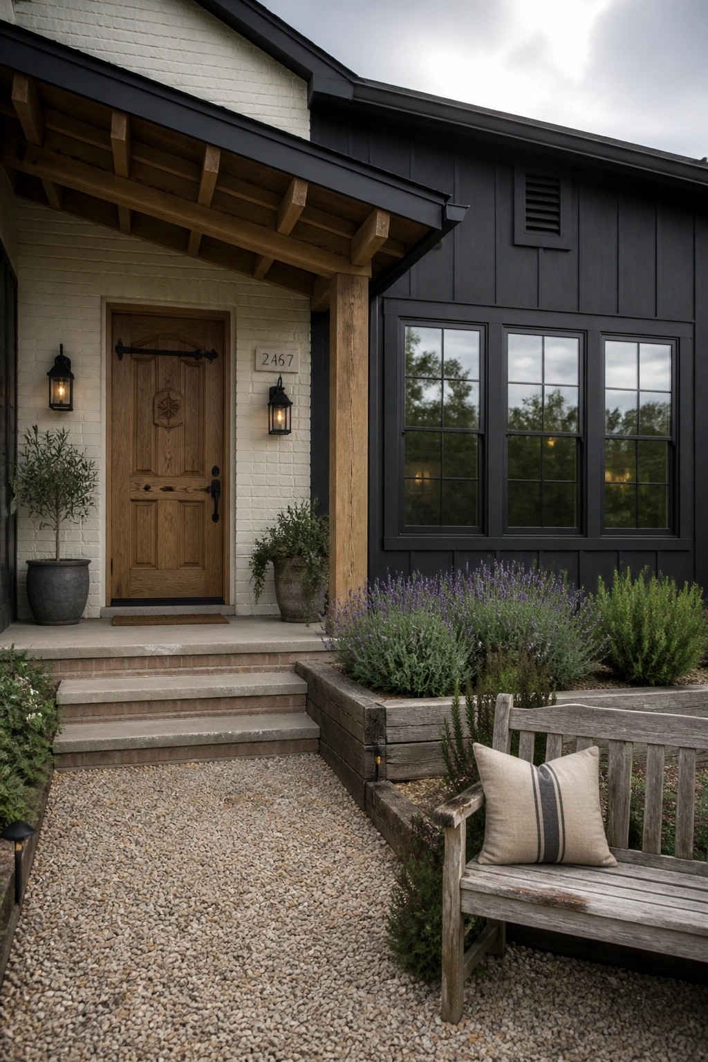 A farmhouse front entry with white brick surrounding a wooden door, black siding on the main wall, and a gravel path leading to steps with a wooden bench nearby.