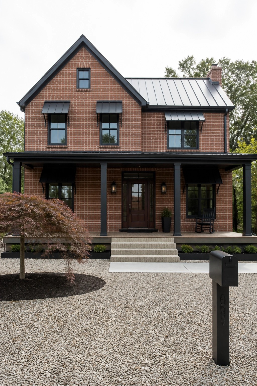 A two-story red brick farmhouse with a dark metal roof, black window awnings, black porch columns, and a gravel front yard.