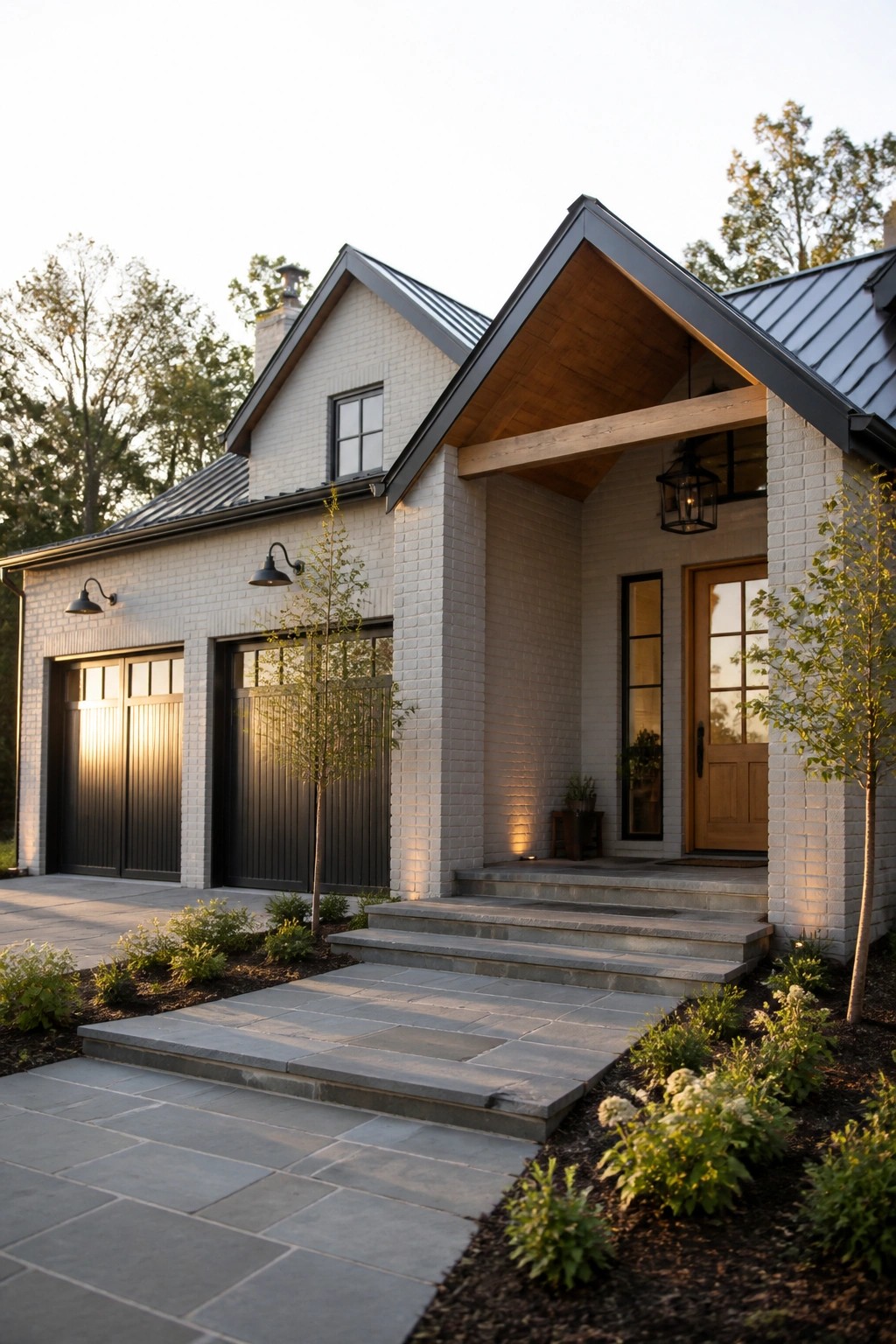 A white brick farmhouse exterior with a dark metal roof, black garage doors, a gabled wood entry porch, and stone steps leading to a wooden front door.