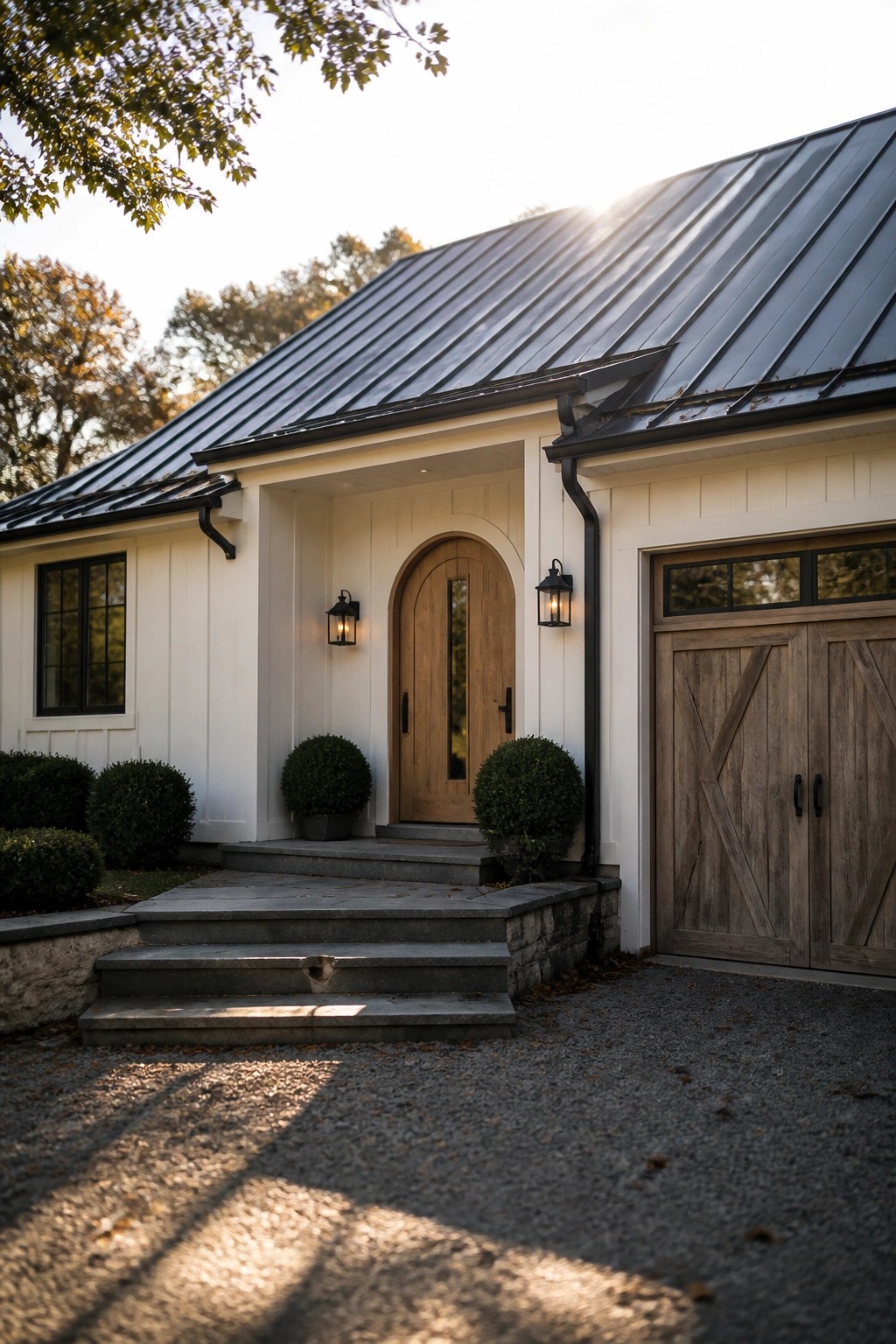 A white farmhouse exterior with an arched wooden front door, black lanterns on either side, stone steps, and a dark metal roof.