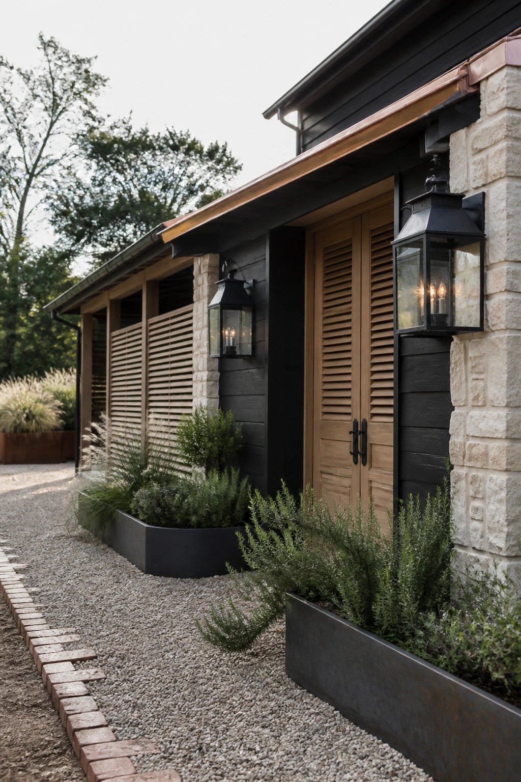 A side view of a farmhouse exterior featuring black horizontal siding, light stone pillars, wooden double doors with shutters, and black metal lanterns mounted on the wall beside a gravel path.