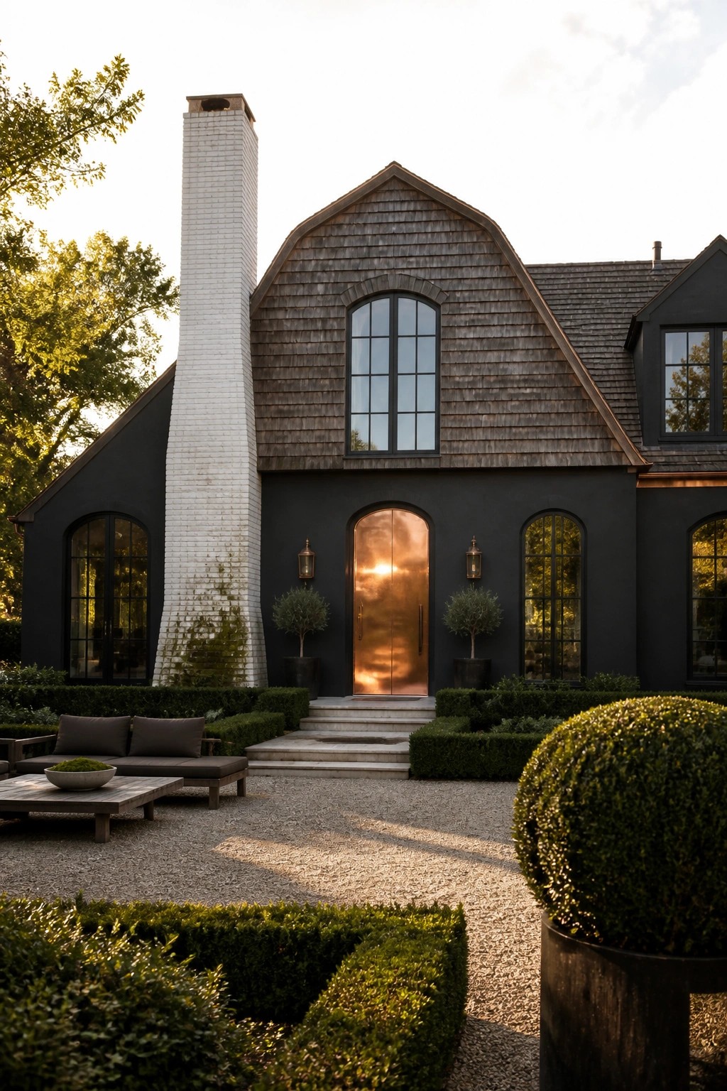 A dark gray farmhouse with a tall white brick chimney, arched copper front door, and gravel courtyard with hedges.