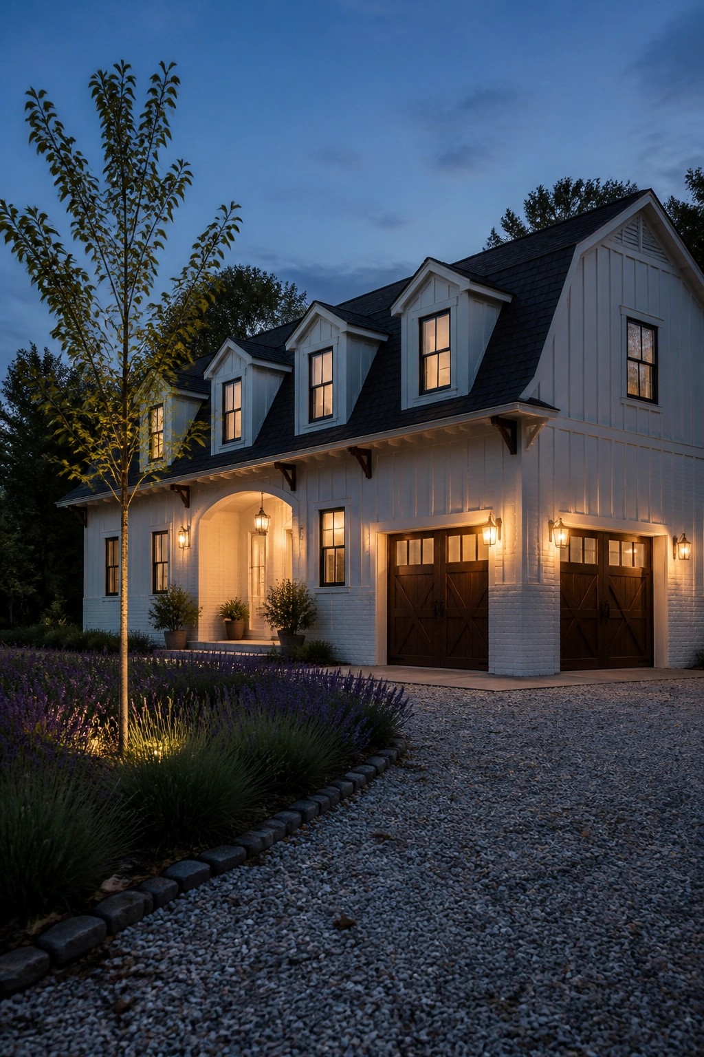 White barn-style house with lit dormers, garages, lavender garden, and gravel drive at dusk.