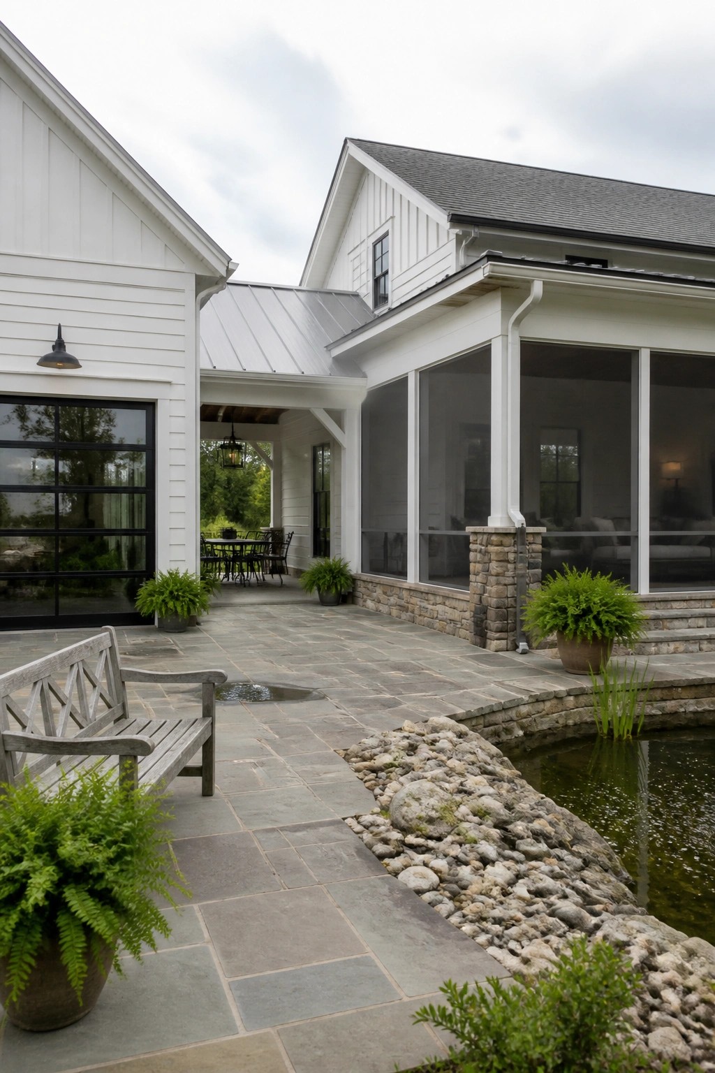 A white farmhouse with a stone porch base, metal roof, black-framed windows, and a stone patio beside a small pond.