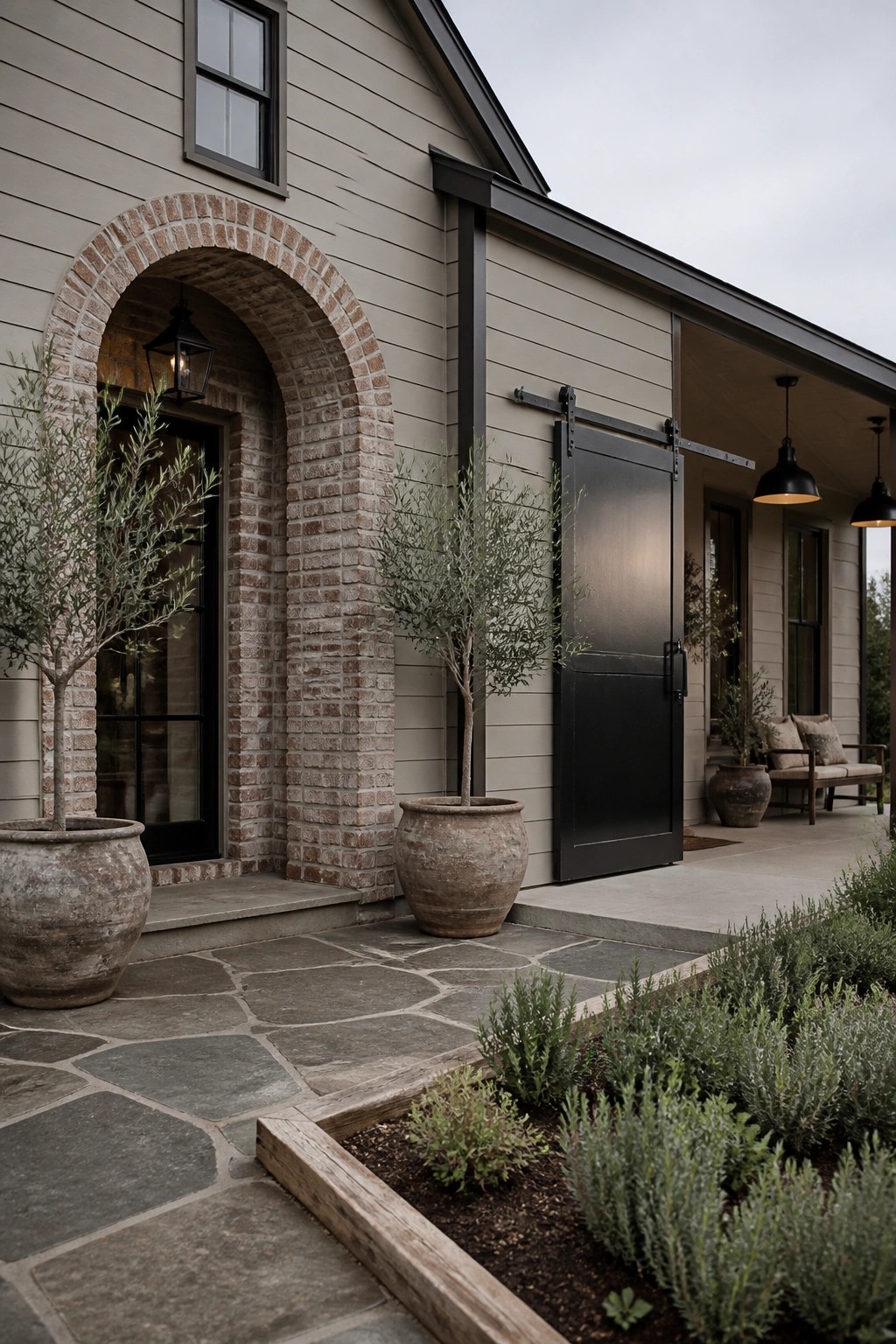 A house exterior features light beige horizontal siding, a brick arched doorway with a black door, a black sliding barn door, and large potted trees on a stone walkway.