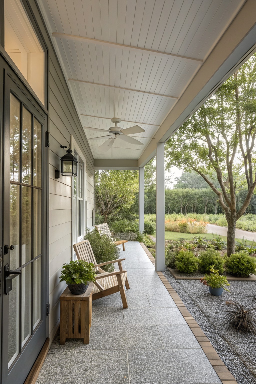 A covered porch with stone tile flooring, wooden armchairs, a ceiling fan, potted plants, and a view of a garden and gravel path.