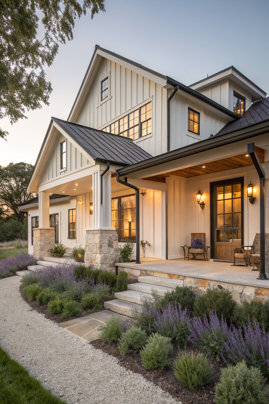 A white farmhouse exterior with a covered porch supported by stone columns, black trim, and purple flowering plants along the front.