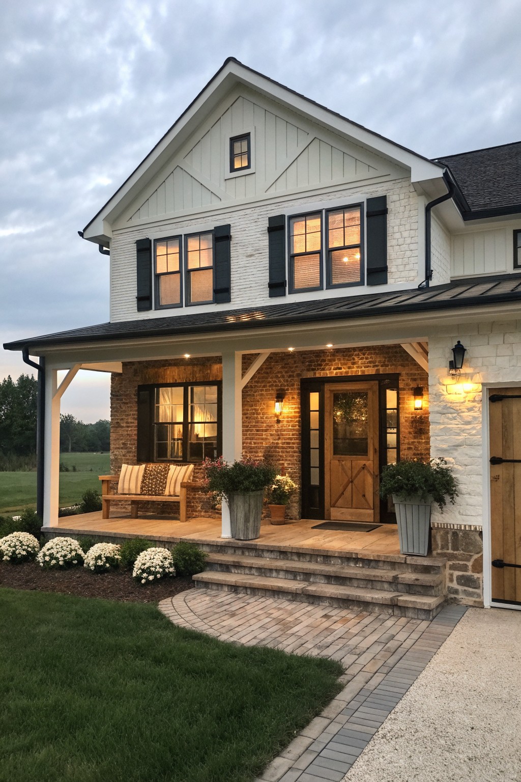 White two-story farmhouse with brick porch, wooden bench, potted plants, and lit windows