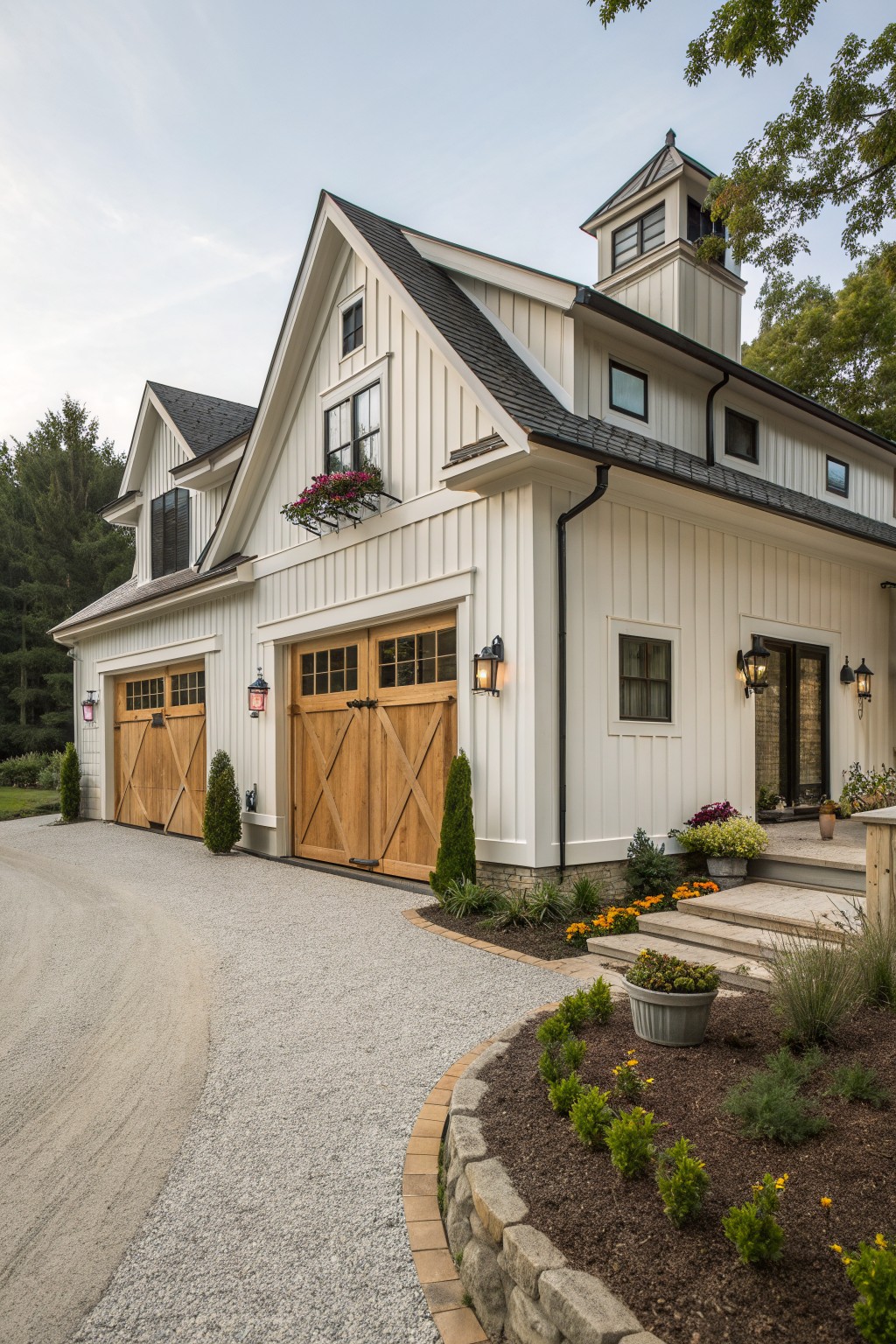 A white two-story house with wooden barn-style garage doors, a cupola, and a gravel driveway bordered by garden beds.
