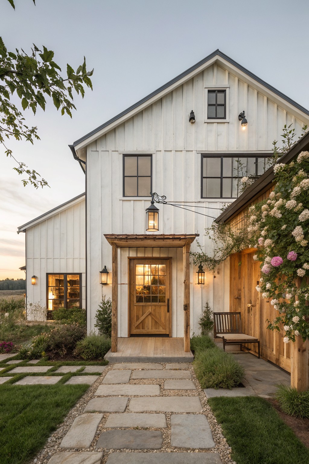 White barn house with wooden door, stone path, lanterns, and blooming vines at dusk.