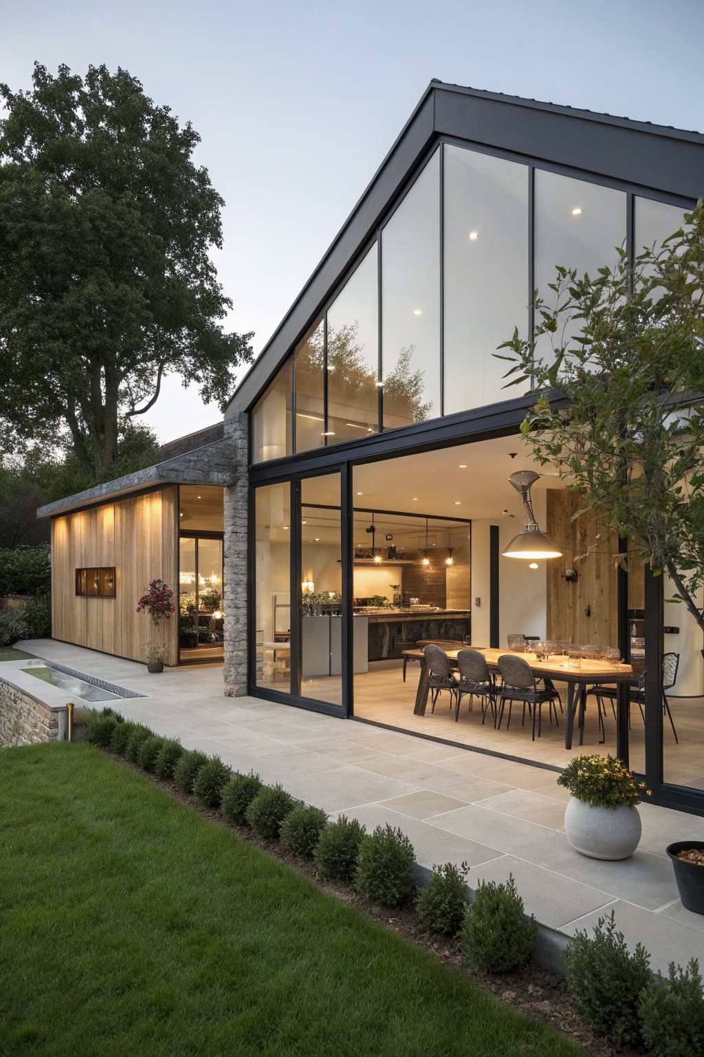 A modern barn-style house exterior featuring wood siding, stone walls, large black-framed glass windows and doors, and an adjacent stone patio with outdoor dining furniture.