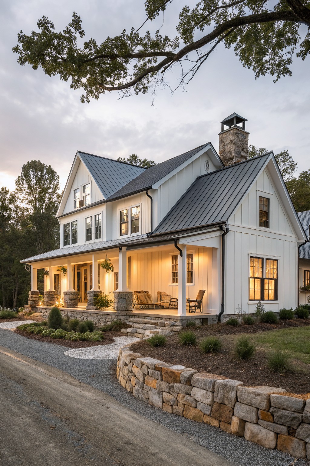 White modern farmhouse with dark metal roof, stone chimney, and lit porch at dusk