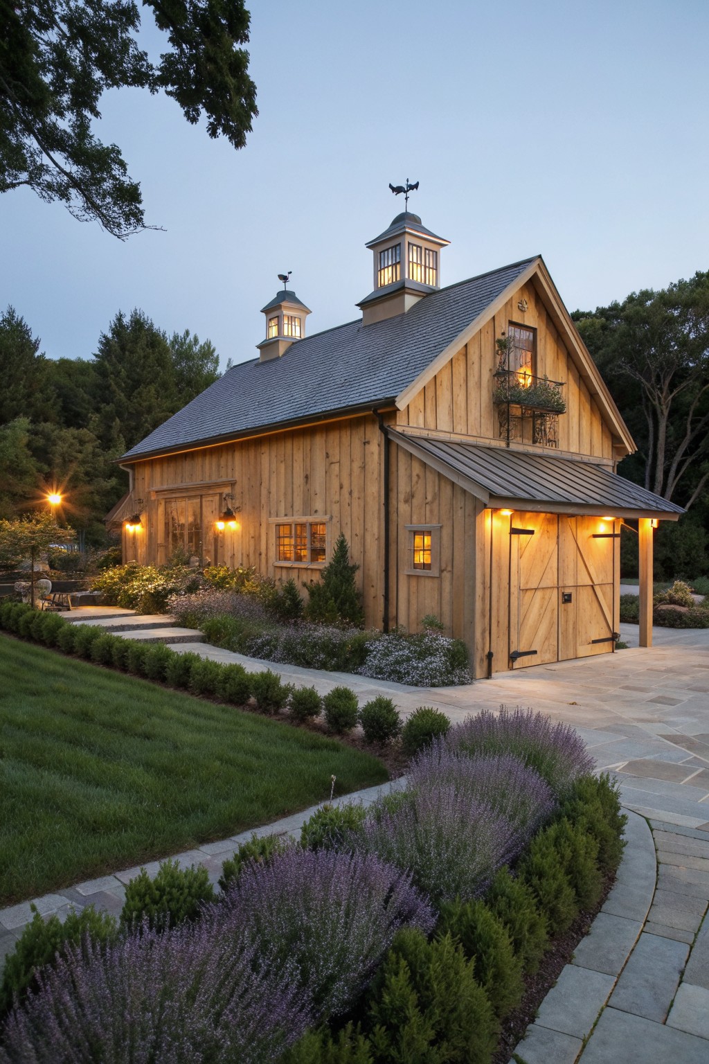 Wooden barn with cupolas and balcony illuminated at dusk amid purple flowers and greenery.