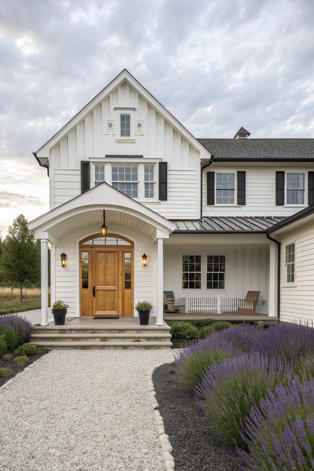A white two-story farmhouse with a wooden front door centered under an arched porch, black shutters on the windows, and lavender plantings along a gravel path.