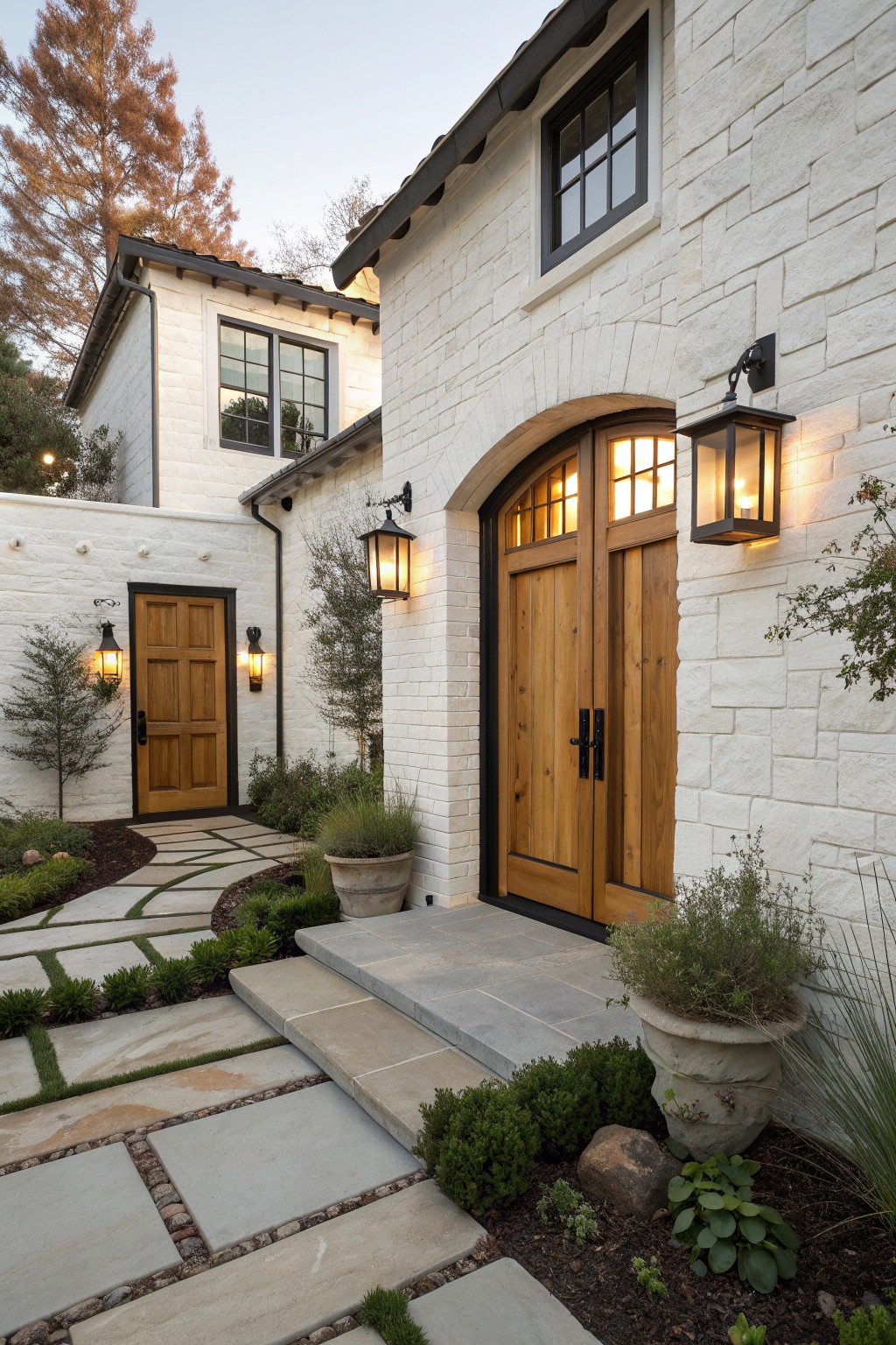 Front view of a white stone house with an arched wooden double door entry, side wooden door, black-framed windows, and lit outdoor lanterns along a stone pathway.