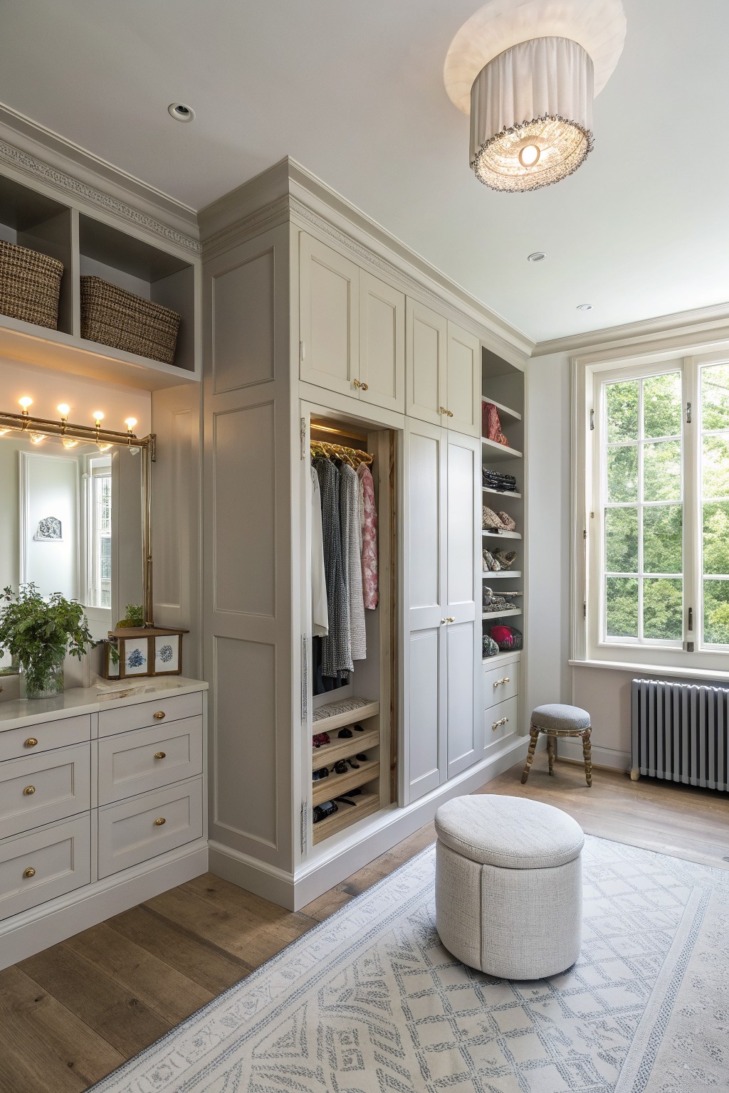 A bright walk-in closet with cream built-in cabinets, open shelving, a vanity with mirror and lights, and two round upholstered ottomans on a patterned rug.