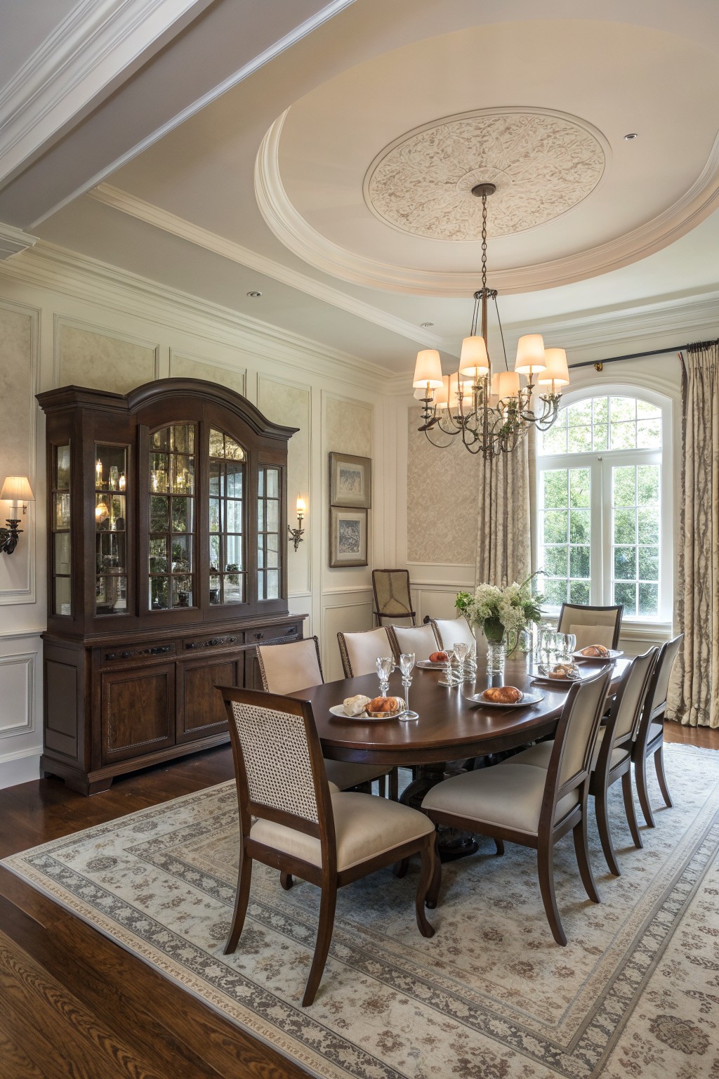 A dining room with a large dark wood hutch along the left wall, a round table set with plates and glasses on a patterned rug, and a chandelier beneath a decorative ceiling.