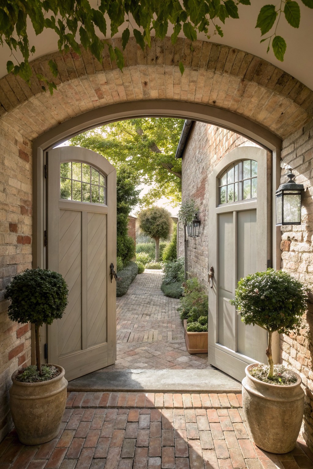 A brick arched doorway with open double doors leads to a paved garden path flanked by two large potted shrubs.
