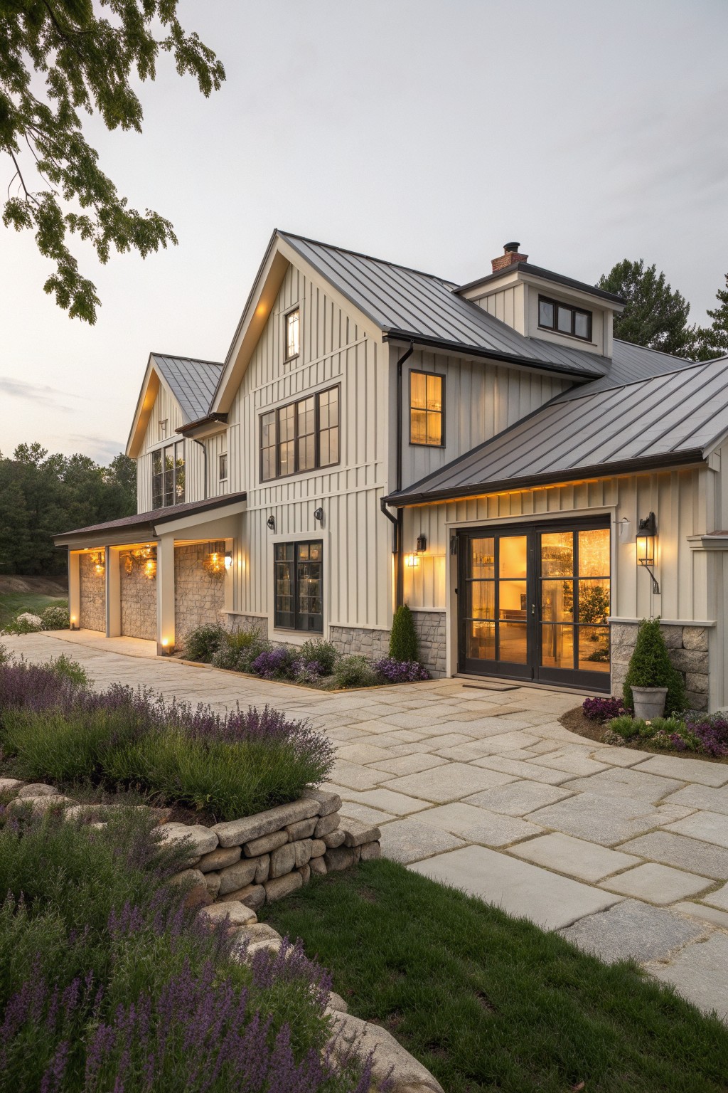Modern white farmhouse with metal roof, stone accents, lit windows, and stone driveway at dusk.