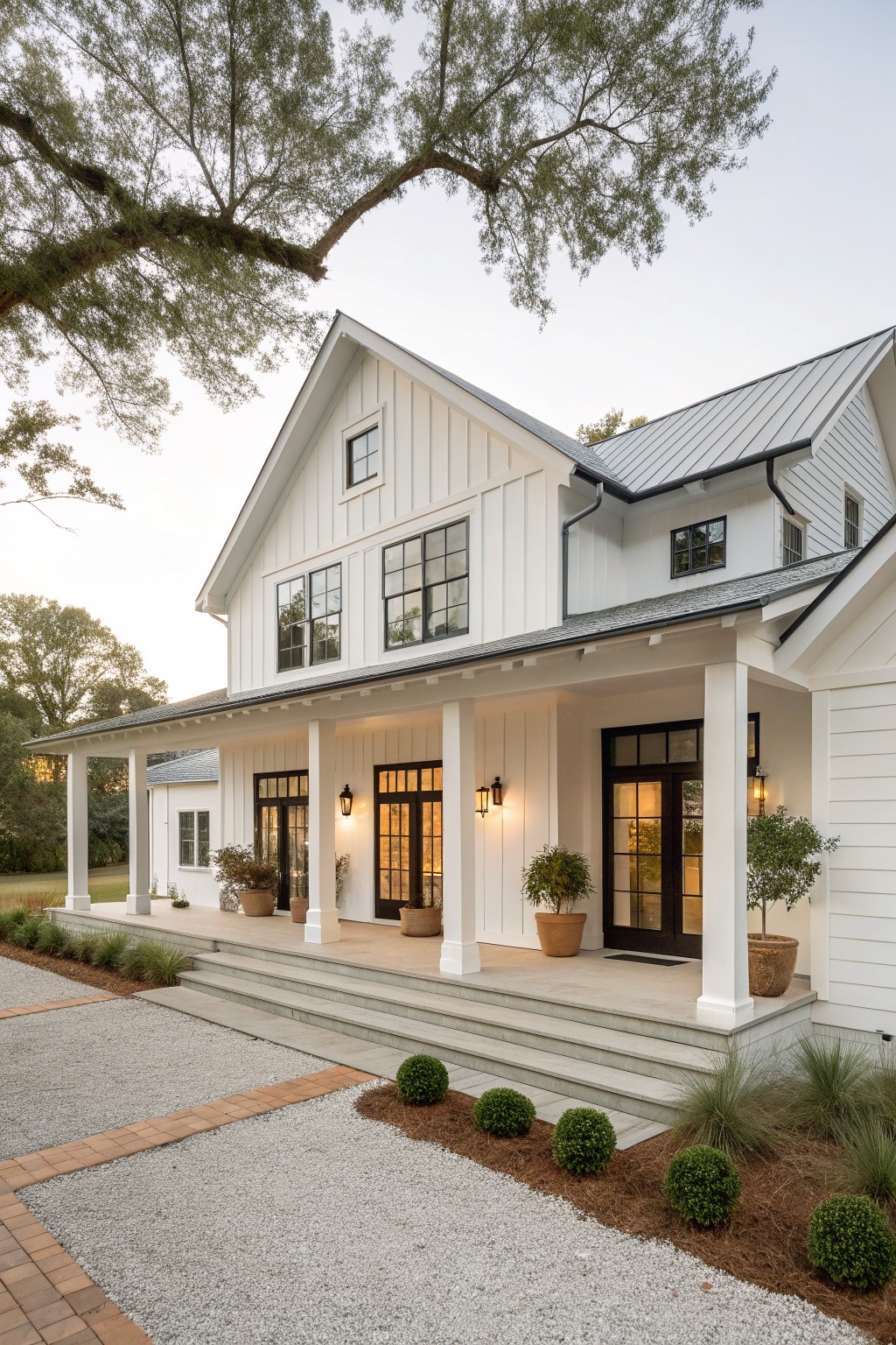 A white two-story farmhouse exterior with black-framed windows and doors, a covered front porch supported by white columns, and a gravel driveway leading to the steps.