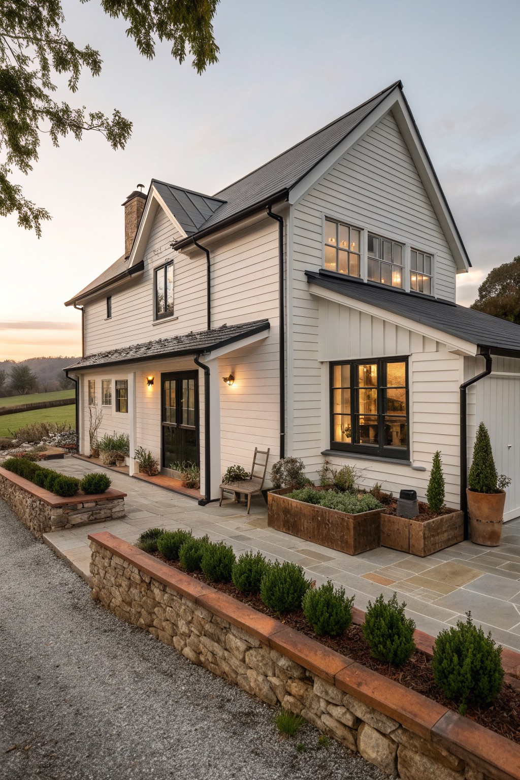 White house with dark trim, lit windows, wooden planters on stone patio at dusk