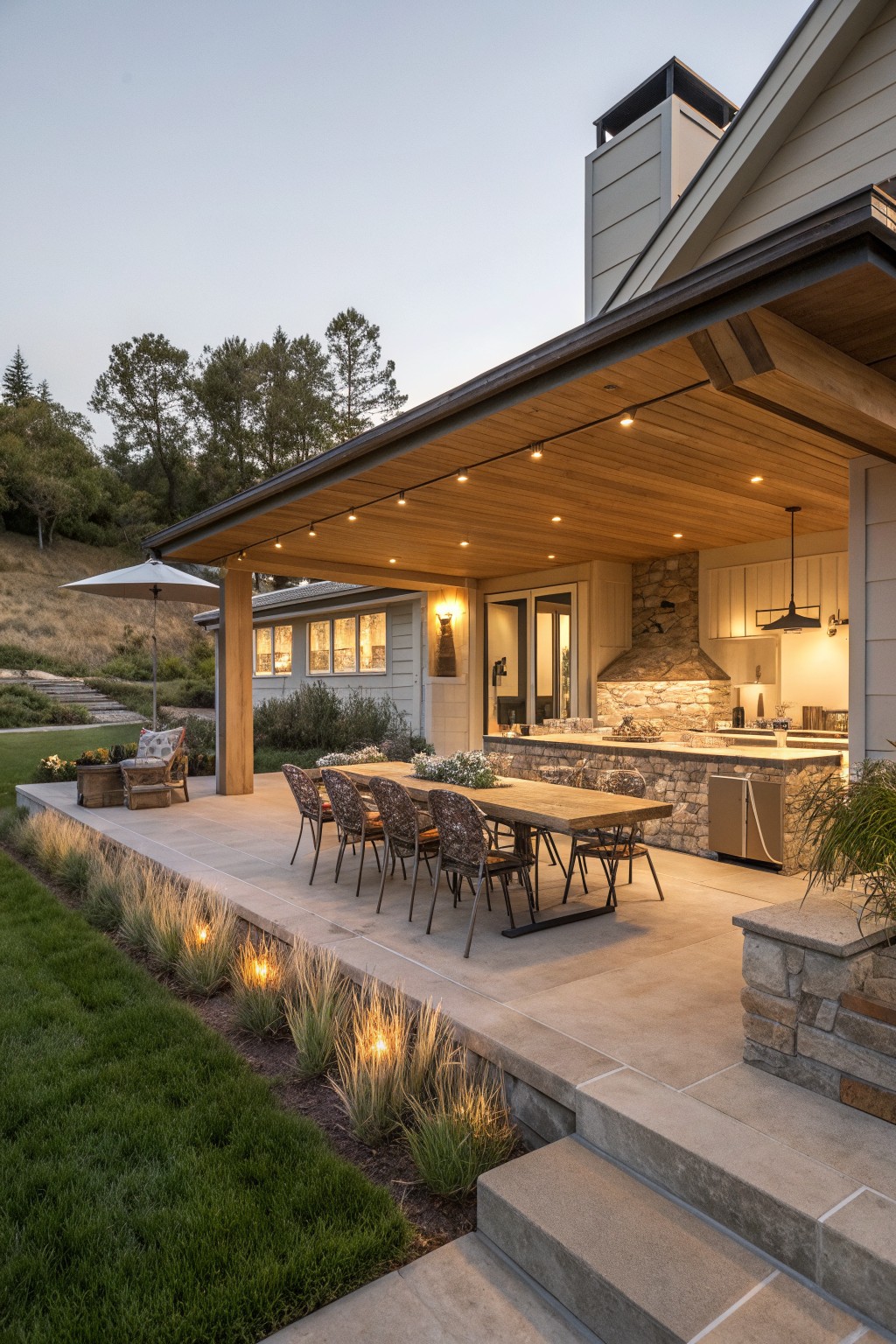 A covered outdoor patio attached to a house features a long wooden dining table with chairs and a stone outdoor kitchen with bar seating under a wood ceiling with string lights.