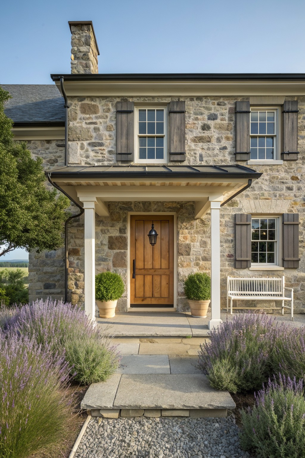 Stone farmhouse exterior with a centered wooden door under a covered porch supported by white columns, potted shrubs beside the door, and lavender plantings along the stone steps.