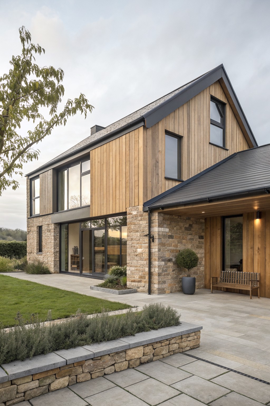 Modern farmhouse exterior with a stone lower section, vertical wood cladding on the upper walls, dark roof trim, and a paved entry area with a bench.