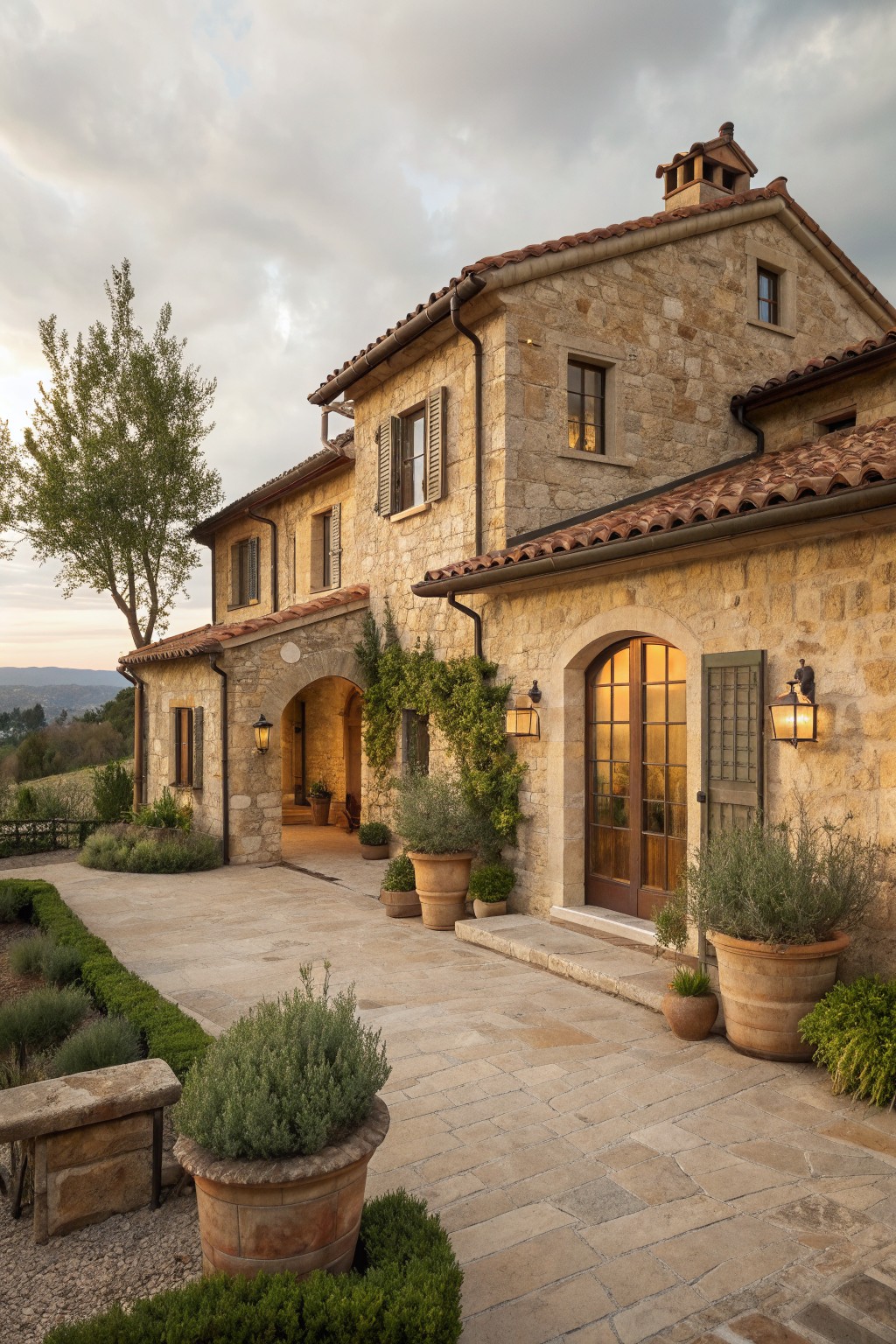 A two-story stone farmhouse with terracotta roof tiles, wooden doors and shutters, arched entryways, and large terracotta pots placed along the paved courtyard.