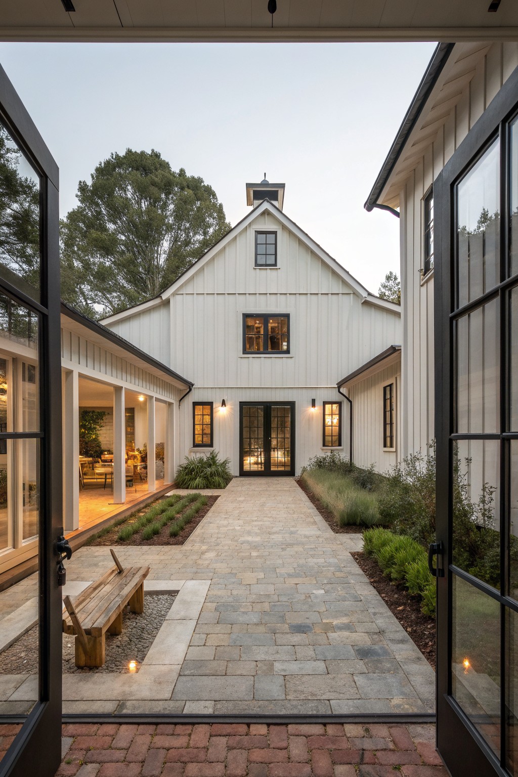 A white barn-style house viewed from an open doorway, with black-framed windows and doors, a central stone path, and simple landscaping on both sides.