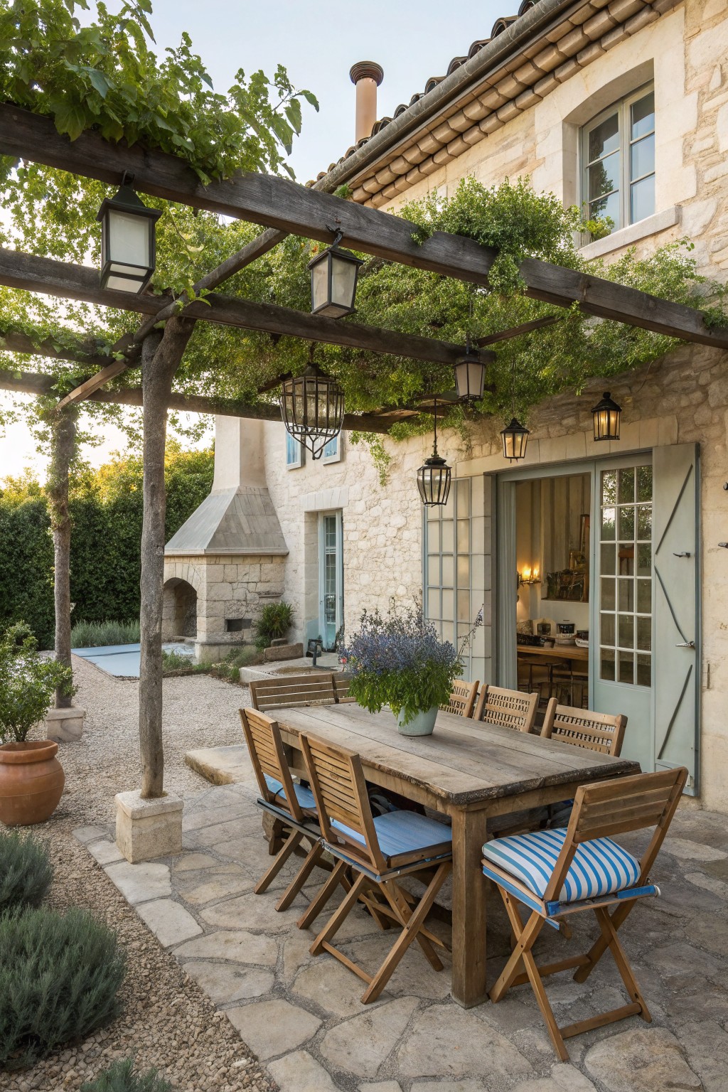 Stone farmhouse with wooden pergola over a long outdoor dining table and chairs on a paved patio, lanterns hanging from the beams and vines growing across the structure.