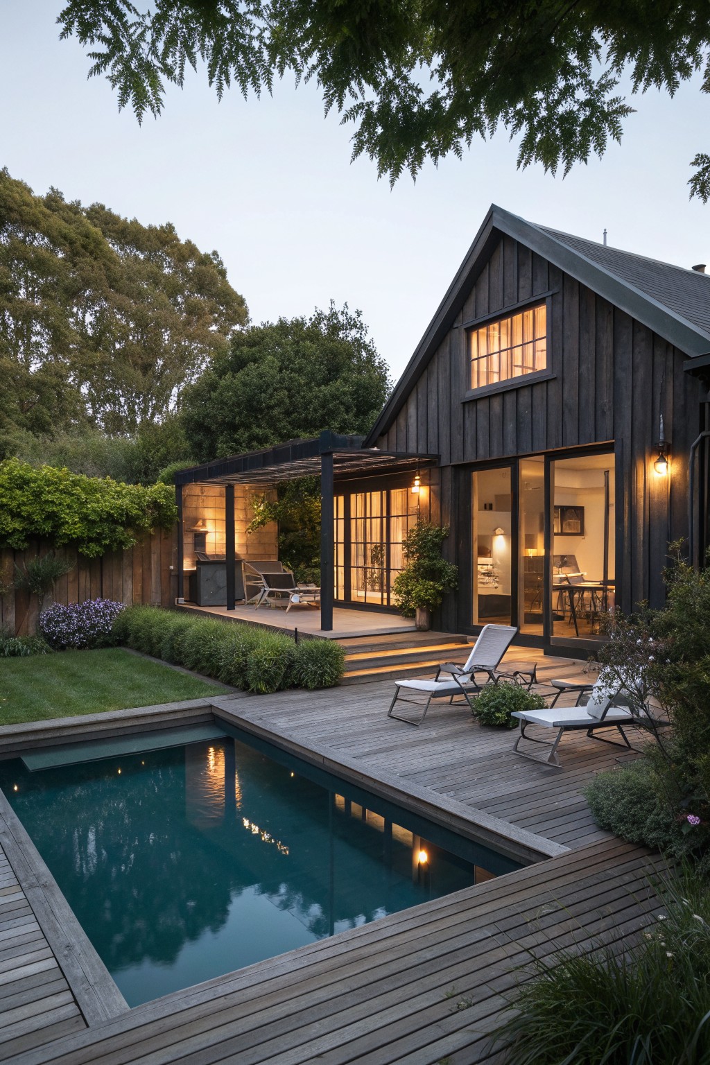 A dark wooden cabin exterior with attached deck, rectangular pool, and lounge chairs surrounded by trees and greenery at dusk.