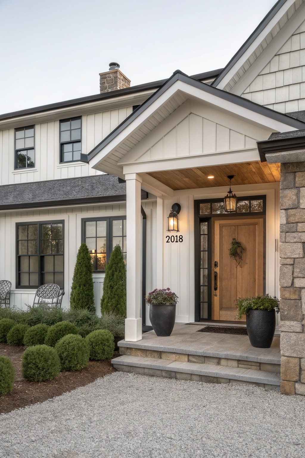 A front view of a modern farmhouse with white vertical siding, black trim, a gabled porch with white columns, a wooden door, and stone on the chimney and right side wall.