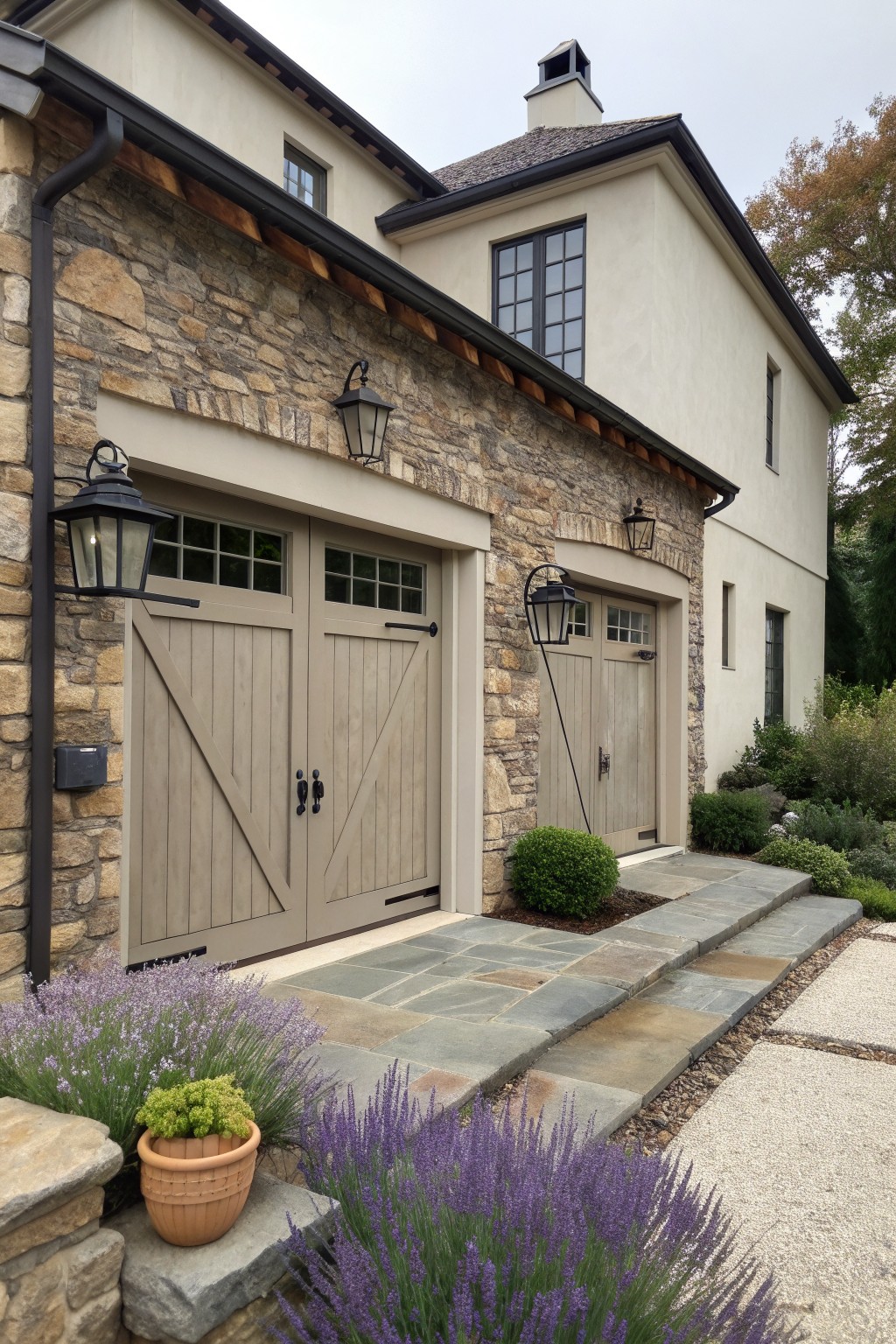 A two-story house exterior with stone on the left side and stucco on the right, featuring two light wood garage doors and black lanterns.