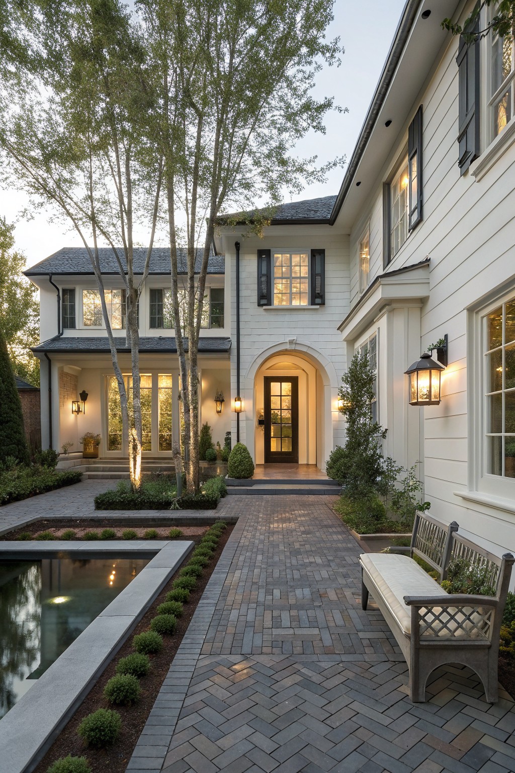 White house with arched entry, brick path, pond, benches, and lit windows at dusk.