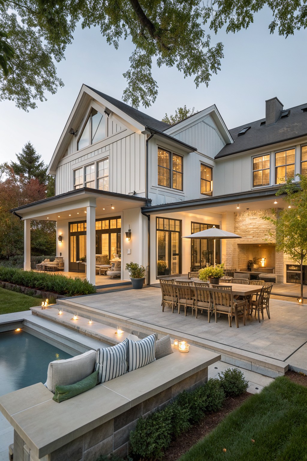 A white modern farmhouse with a large covered patio holding a long wooden dining table, a rectangular pool with built-in bench seating, and a stone outdoor fireplace.